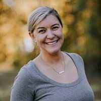 A smiling woman wearing a gray shirt and necklace, outdoors, with her hair pulled back.