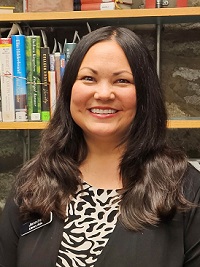 A smiling woman with long dark hair stands in front of a bookshelf.